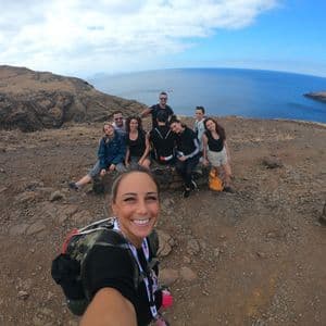 Une femme souriante prend un selfie avec son groupe de voyage WeRoad sur une falaise rocheuse surplombant la mer sous un ciel bleu nuageux.