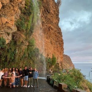Un groupe WeRoad en voyage posant sur une route côtière, sous une cascade s'écoulant d'une falaise rocheuse.