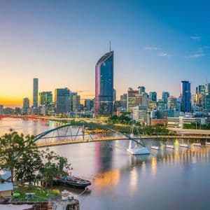 Un panorama urbain moderne avec des gratte-ciel illuminés, se reflétant dans une large rivière au coucher du soleil, et un pont piétonnier au premier plan.
