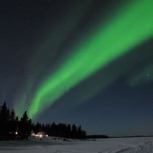 Un'aurora boreale verde illumina un cielo notturno stellato su un paesaggio innevato con alberi e piccole baite.