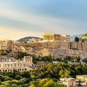 Antike griechische Ruinen auf einer felsigen Akropolis, von goldenem Licht beleuchtet, überblicken eine moderne Stadt und dichte grüne Bäume.