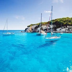 Mehrere Segelboote liegen in einer Bucht vor Anker, mit klarem, hellem, türkisfarbenem Wasser neben einer felsigen, baumbestandenen Klippe.