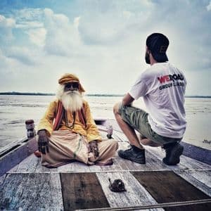 Un líder de grupo de WeRoad y un hombre mayor con barba blanca se sientan en la proa de un barco de madera en un río ancho.