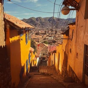 Eine schmale, sonnenbeschienene Steingasse mit Treppen führt zwischen bunten Gebäuden hinab und gibt den Blick auf eine Stadt und Berge frei.