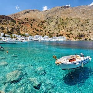 Ein kleines Boot liegt in klarem, türkisfarbenem Wasser in einer Bucht mit einem weiß getünchten Dorf am Fuße eines Berges vor Anker.