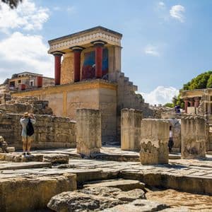 A person wearing a hat photographs the ancient stone ruins of a palace with red columns under a partly cloudy sky.