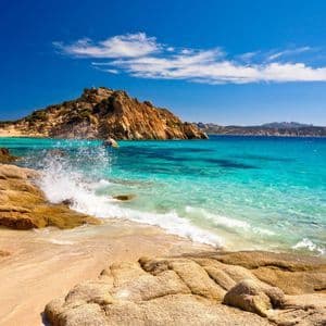 Una ola rompe contra grandes rocas en una playa de arena con agua clara y turquesa bajo un cielo azul brillante con algunas nubes.