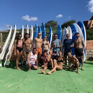A WeRoad group trip in swimwear poses for a photo in front of a rack of upright surfboards on a sunny day.