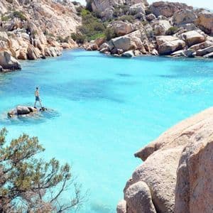 Una persona en bañador de pie sobre una pequeña roca en una cala con agua turquesa cristalina, rodeada de acantilados rocosos.