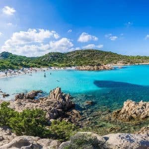 Menschen schwimmen in türkisfarbenem Wasser und sonnen sich an einer weißen Sandstrandbucht, umgeben von felsigen grünen Hügeln unter einem strahlend blauen Himmel.