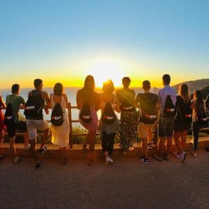 A WeRoad group trip seen from behind, watching a golden sunset over the sea from a coastal viewpoint.