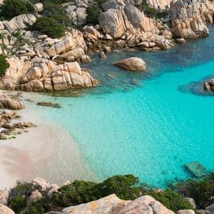 Una caletta sabbiosa e appartata con acqua turchese cristallina, circondata da grandi rocce e arbusti verdi, vista dall'alto.