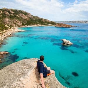 Un uomo in camicia blu siede su una grande roccia che domina una baia turchese e cristallina con una costa rocciosa.