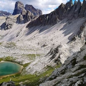 Vista dall'alto di un lago alpino turchese in una vasta valle rocciosa con cime frastagliate sotto un cielo parzialmente nuvoloso.
