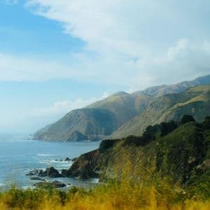 Une vue sur une côte accidentée, où des montagnes vertes descendent vers l'océan bleu sous un ciel partiellement nuageux, avec un pont au loin.
