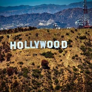 Le panneau blanc Hollywood se dresse sur une colline sèche et broussailleuse, avec une tour de communication à proximité et des montagnes en arrière-plan.
