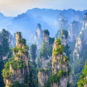 De hauts et minces piliers rocheux coiffés de végétation verte s'élèvent d'une forêt dense, avec des montagnes brumeuses en arrière-plan.