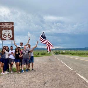 Eine WeRoad-Gruppe posiert an einem historischen US Route 66 Schild in Arizona, wobei eine Person eine amerikanische Flagge neben einer ländlichen Straße hält.