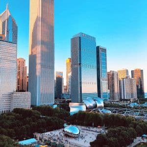 Vue aérienne d'un parc urbain présentant une grande sculpture réfléchissante en forme de haricot sur une place, entourée de gratte-ciel modernes sous un ciel bleu clair.