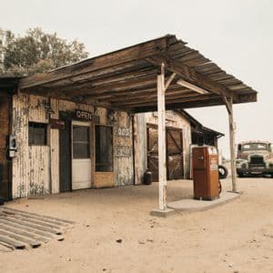 Une station-service rustique avec des murs en tôle ondulée, une pompe à essence orange vintage et un vieux camion garé sur un terrain sablonneux.