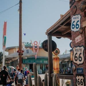 Une scène de rue ensoleillée avec des panneaux Route 66 sur un bâtiment en briques, des gens marchant près de cactus et un panneau V8 rétro en arrière-plan.