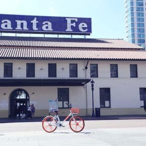 An orange and white bicycle is parked on a city sidewalk in front of the historic Santa Fe Depot building on a sunny day.