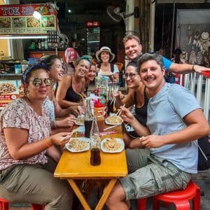 Eine lächelnde WeRoad-Reisegruppe sitzt gemeinsam an einem Holztisch und isst Nudeln von einem Streetfood-Stand.