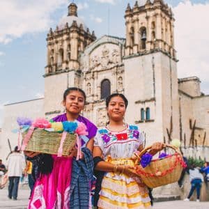 Dos mujeres jóvenes con coloridos vestidos tradicionales sostienen cestas de flores de papel, posando frente a una gran iglesia de piedra.