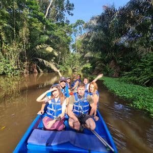 Un voyage de groupe WeRoad navigue sur une rivière de jungle dans un bateau bleu, au milieu d'un feuillage tropical dense.