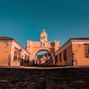 Vue en contre-plongée d'une arche jaune surmontée d'une tour d'horloge, enjambant une rue pavée entre deux bâtiments, sous un ciel bleu clair.