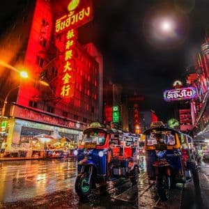 Dos tuk-tuks coloridos estacionados en una calle urbana mojada por la noche, iluminados por luminosos letreros de neón rojos en los edificios.