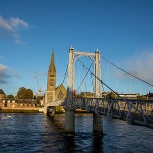 Eine weiße Hängebrücke überspannt einen Fluss mit historischen Kirchen und Gebäuden am Ufer unter blauem Himmel.