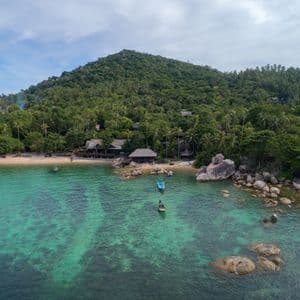 Eine Luftaufnahme einer tropischen Bucht mit klarem türkisfarbenem Wasser, kleinen Booten, einem Sandstrand und einem üppig grünen, baumbestandenen Hügel.