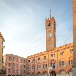 Ein sonnenbeschienener, roter Backsteinglockenturm und ein angrenzendes Bogengebäude stehen auf einem historischen Marktplatz unter einem hellblauen Himmel.