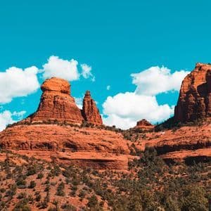 Grandi formazioni rocciose rosse si ergono da un paesaggio di macchia verde sotto un cielo azzurro brillante con nuvole bianche.