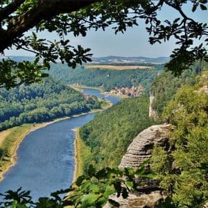 Ein Blick aus der Vogelperspektive auf einen breiten Fluss, der sich durch ein grünes, bewaldetes Tal, vorbei an einem kleinen Dorf und felsigen Klippen schlängelt.