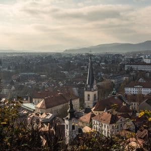 Eine Stadtansicht mit einem markanten Kirchturm und rotgedeckten Gebäuden unter einem bewölkten Himmel, mit Bergen in der Ferne.