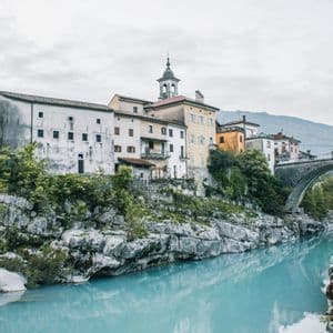 Eine Stadt mit Glockenturm liegt auf einer felsigen Klippe über einem helltürkisen Fluss und einer Steinbrücke.