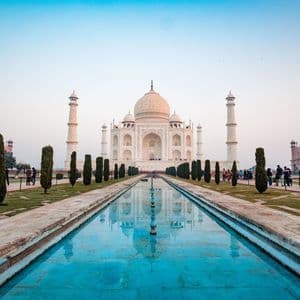 Il Taj Mahal si riflette in una lunga piscina fiancheggiata da giardini con cipressi, con visitatori che passeggiano sui sentieri.
