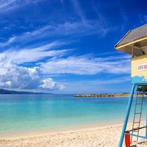 Una torre de salvavidas amarilla y azul se alza en una playa de arena junto a aguas tranquilas de color turquesa bajo un cielo parcialmente nublado.