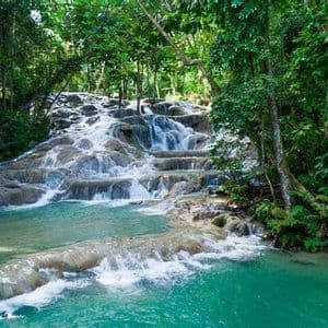 Una cascada escalonada fluye sobre rocas pulidas hacia una poza turquesa, rodeada por un frondoso bosque tropical.
