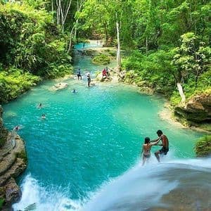 Un grupo de WeRoad disfruta de una cascada selvática de color turquesa, con algunos bañándose abajo y dos de pie al borde del salto de agua.