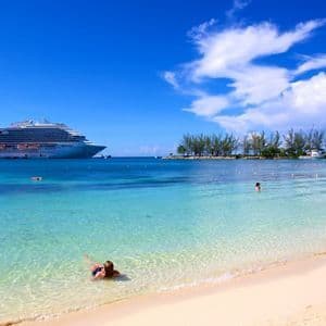 Un crucero anclado en aguas turquesas cerca de una playa de arena con gente nadando bajo un cielo azul y soleado.