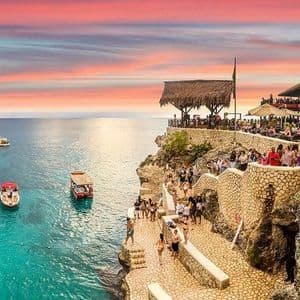 La gente se reúne en un acantilado de piedra con un restaurante, observando barcos en el agua turquesa durante una colorida puesta de sol.