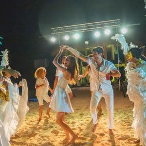 Un viaje en grupo de WeRoad bailando en la arena por la noche durante una fiesta en la playa con artistas en elaborados disfraces de plumas.