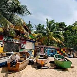 Varios botes pequeños y coloridos descansan en una playa de arena frente a una cabaña rústica y exuberantes palmeras.