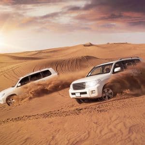 Zwei weiße SUVs fahren bei Sonnenuntergang große Wüstendünen hinunter und wirbeln dabei Sand auf.
