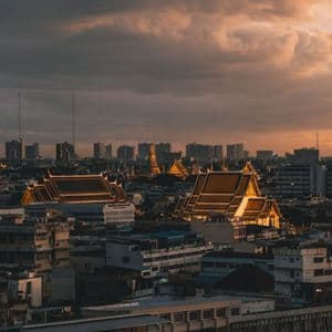 Beleuchtete Tempeldächer heben sich bei Sonnenuntergang unter bewölktem Himmel von einer dichten Stadtlandschaft mit moderner Skyline ab.