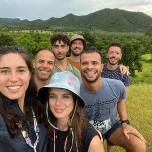 Un voyage en groupe WeRoad de sept personnes souriant pour un selfie dans un champ herbeux, avec en toile de fond des collines et des arbres verdoyants.