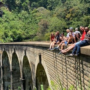 Un gruppo WeRoad in viaggio, seduto insieme sul bordo di un grande ponte ad arco in pietra, in una lussureggiante valle verde.